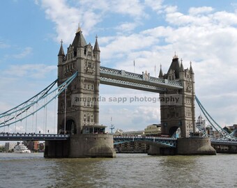 Puede incluir: Una vista del icónico Puente de la Torre en Londres, Inglaterra. El puente está hecho de piedra y metal, y tiene dos torres a cada lado de la pasarela. El puente está sobre una masa de agua, y hay barcos en el agua.