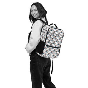 May include: A white backpack with a repeating pattern of spiders in red, blue, and black. The backpack has a black pocket and straps. The person in the image is wearing the backpack.