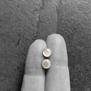 May include: Two round, iridescent stud earrings with silver settings. The earrings are displayed on a grayscale background.