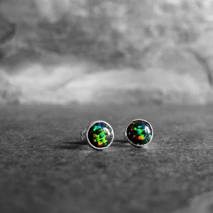 May include: A pair of round stud earrings with a black opal center. The opals display a vibrant array of colors, including green, orange, and blue, set in a silver bezel. The earrings are displayed on a gray surface.