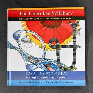 May include: A hardcover book titled "The Cherokee Syllabary" with a colorful illustration of a stylized flower and Cherokee syllabary characters. The cover features text in English and Cherokee, with the author and illustrator's names.