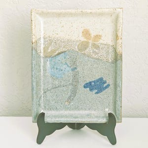 May include: Rectangular ceramic serving platter with a speckled off-white top and a blue and grey patterned bottom. The platter features a floral design and is displayed on a dark green stand. A decorative piece for the home.