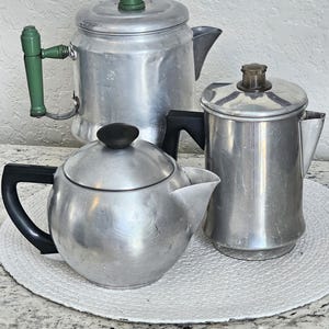 May include: Three silver aluminum coffee pots with black handles. The coffee pots are on a white crocheted placemat.
