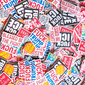 May include: A collection of stickers and buttons with various anti-Trump messages. The stickers feature phrases like "FUCK TRUMP," "ICE," and "Blessed Be The Fight." Colors include red, white, black, and blue.