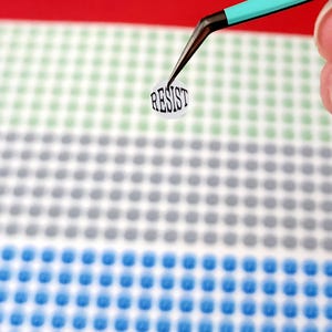 May include: A small, round sticker with the word "RESIST" in black, held by teal tweezers. The sticker is on a background of small, colorful dots in rows of blue, gray, and green, with a red stripe at the top.