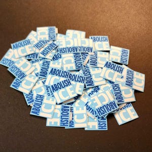 May include: A pile of small, square stickers with the words "ABOLISH ICE" in white block letters on a light blue background. The stickers are scattered on a dark surface.