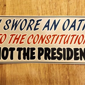 May include: Rectangular sticker with the text "I SWORE AN OATH TO THE CONSTITUTION NOT THE PRESIDENT" in blue, red, and black lettering on a silver background. The sticker is on a wooden surface.