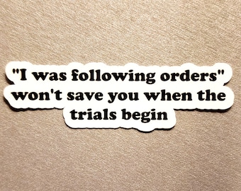 May include: White sticker with black text that reads: "I was following orders" won't save you when the trials begin. The sticker has a white border and is on a tan background.