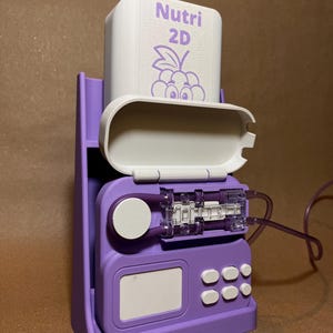 Op de afbeelding: Een 3D-geprint medisch apparaat in paarse en witte tinten. Het apparaat heeft een rechthoekige witte container met het opschrift "Nutri 2D" en een druivenillustratie. Het heeft een transparante kunststof component en diverse knoppen.