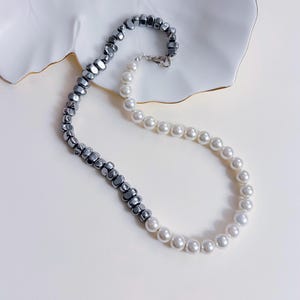 以下が含まれることがあります： ダークグレーの不規則な形のビーズと白い丸い真珠を組み合わせた、ツートーンのネックレス。 ネックレスはシルバーカラーの留め金で留められています。