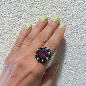 以下が含まれることがあります： 白い真珠のハローで囲まれた、大きな濃いピンクの宝石が付いた銀の指輪です。