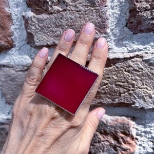 May include: A large, square ring with a deep red, almost magenta, colored stone set in a silver band.