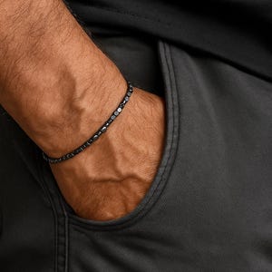 Op de afbeelding: Een donkergrijze kralenarmband met een minimalistisch design. De armband is gemaakt van kleine, ronde kralen en wordt om een pols gedragen. De persoon draagt een zwart shirt en een grijze broek.