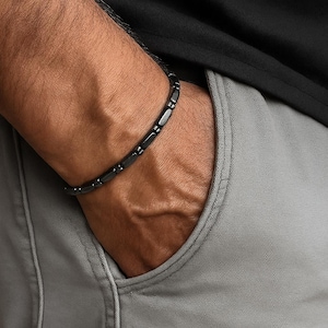 Puede incluir: Una pulsera negra con eslabones rectangulares se lleva en la muñeca. La pulsera está hecha de un material oscuro y contrasta con el tono de la piel. La persona lleva una camisa negra y pantalones grises.