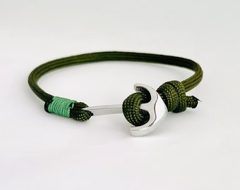 Puede incluir: Una pulsera de paracord verde con un dije de ancla de plata. La pulsera está atada con un nudo y tiene un acento verde y verde claro.
