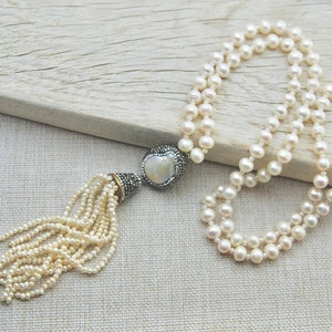 Pode incluir: Um colar longo de p&eacute;rolas brancas com um pingente de cora&ccedil;&atilde;o prateado e branco e um tassel de p&eacute;rolas menores.