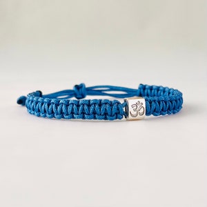 May include: A blue braided bracelet with a silver charm featuring the Om symbol.