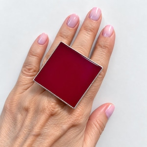 Puede incluir: Un anillo cuadrado grande con una incrustación central plana de color magenta intenso, engastado en un marco de metal fino de tono plateado pulido. El diseño geométrico cubre una parte importante del dedo, mostrando un color sólido y llamativo.