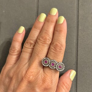 Könnte beinhalten: Ein silberner Ring mit drei kreisförmigen Designs. Jeder Kreis hat einen zentralen roten Stein, der von kleineren weißen Steinen umgeben ist. Der Ring wird an einer Hand mit gelbem Nagellack getragen.