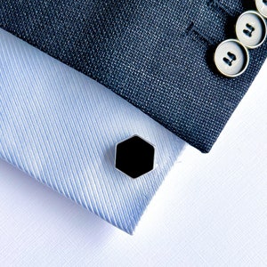 Puede incluir: Un botón de camisa hexagonal negro con borde plateado. El botón de camisa está sobre una tela a rayas azul y blanca.