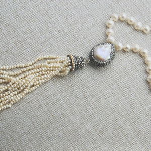 Pode incluir: Um colar de p&eacute;rolas brancas com um pingente de p&eacute;rola grande e irregular e um tassel de p&eacute;rolas menores. O pingente &eacute; engastado em um metal prateado com um acabamento brilhante.