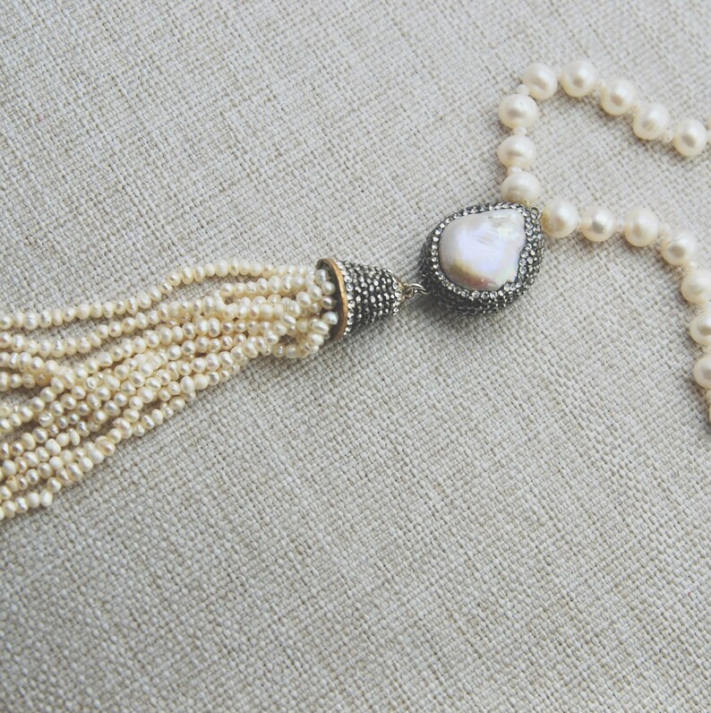 Pode incluir: Um colar de p&eacute;rolas brancas com um pingente de p&eacute;rola grande e irregular e um tassel de p&eacute;rolas menores. O pingente &eacute; engastado em um metal prateado com um acabamento brilhante.