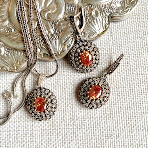 May include: A silver necklace and earrings set with a round pendant and matching earrings. Each piece features a large orange gemstone surrounded by smaller clear gemstones.