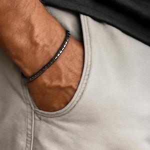Può includere: Un braccialetto grigio scuro composto da piccole perline rettangolari è indossato su un polso. Il braccialetto ha un design semplice e minimalista. La persona indossa una maglietta nera e pantaloncini grigio chiaro.