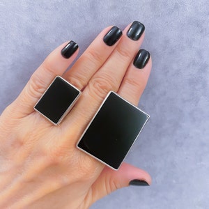 May include: Two black rectangular rings with silver settings. The rings are worn on a hand with black nail polish.