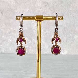 Peut inclure: Une paire de boucles d'oreilles pendantes avec une finition argent et or. Les boucles d'oreilles présentent une grande pierre précieuse rose entourée de petites pierres précieuses claires. Les boucles d'oreilles sont suspendues à un support en or.
