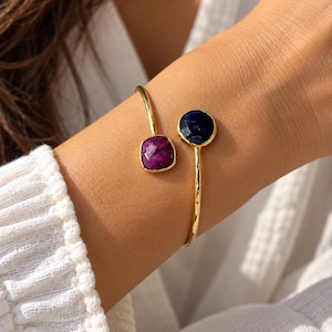 Op de afbeelding: Een goudkleurige open armband met twee gefacetteerde edelstenen. Het ene uiteinde bevat een vierkante, diep magenta steen, terwijl het andere uiteinde een ronde, donkerblauwe steen heeft. De metalen band heeft een gepolijste afwerking met textuur.
