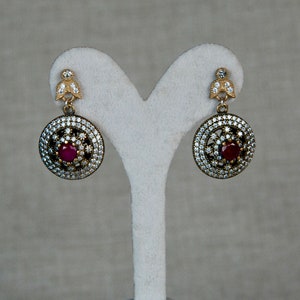 May include: A pair of silver earrings with a red gemstone center and a circle of smaller white gemstones around it. The earrings are hanging from a small gold-colored piece.
