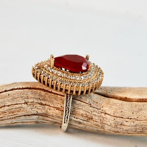 May include: A gold ring with a large red gemstone surrounded by smaller clear gemstones.