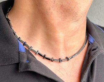 May include: A necklace composed of small, dark, metallic-looking cylindrical beads arranged in a repeating pattern with short, perpendicular bar-shaped spacers between them, resting against the neck.