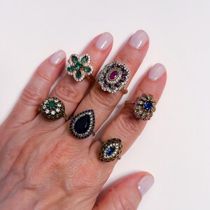 Peut inclure: Un mannequin de main portant six bagues en argent avec des pierres précieuses colorées. Les bagues présentent une variété de motifs, notamment une fleur, une larme et une étoile. Les pierres précieuses sont vertes, bleues, rouges et noires.