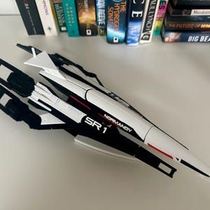 Puede incluir: Maqueta de la nave espacial SR1 Normandy, predominantemente blanca con detalles negros y rojos. El texto "NORMANDY SR 1" es visible en el lateral. Un artículo de colección, probablemente de una franquicia de ciencia ficción.