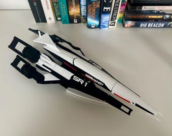 May include: A model of the SR1 Normandy spaceship, predominantly white with black accents and red detailing. The text "NORMANDY SR 1" is visible on the side. The model is a collectible item, likely from a science fiction franchise.