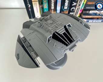 May include: A gray, detailed model of a spaceship with a circular base. The model features intricate paneling, vents, and a central cockpit area. The design includes angled wings and a geometric emblem. Books are visible in the background.