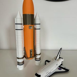以下が含まれることがあります： おもちゃのスペースシャトルとロケット。ロケットはオレンジと白で、茶色のアクセントが付いています。スペースシャトルは白と黒のアクセントで、「United States」の文字が側面に印刷されています。NASAのロゴも表示されています。