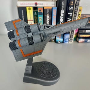 May include: A grey model aeroplane with orange detailing, mounted on a grey stand. The model features detailed engine components and wing designs. The stand has a circular base with an emblem. The model is displayed in front of a bookshelf.