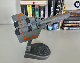 May include: A gray model airplane with orange accents, mounted on a gray stand. The model features detailed engine components and wing designs. The stand has a circular base with an emblem. The model is displayed in front of a bookshelf.