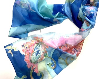Puede incluir: Una bufanda de seda azul y verde con un diseño floral. La bufanda está pintada a mano con flores rosas, naranjas y amarillas. La bufanda está doblada por la mitad y drapeada sobre una superficie blanca.