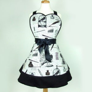 May include: A black and white heart-shaped apron with a vintage-inspired design featuring birds, cages, inkwells, and writing implements. The apron has a black ribbon tie and a black ruffle trim.