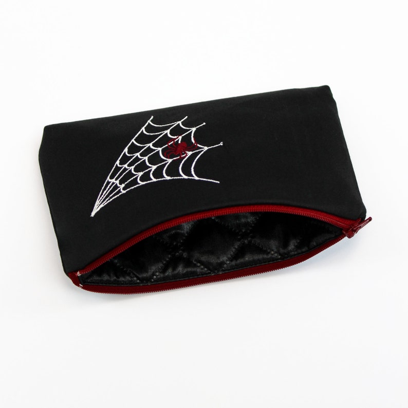 May include: Black zippered pouch with a red zipper and a white embroidered spider web with a red spider in the center.