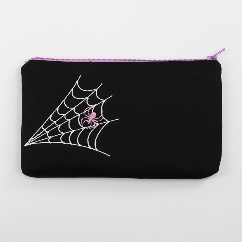 May include: Black zippered pouch with a white embroidered spider web and a pink spider in the center.