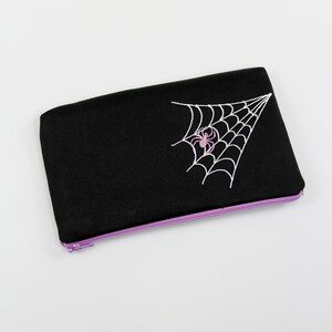 May include: Black zippered pouch with a white embroidered spider web and a pink spider in the center.