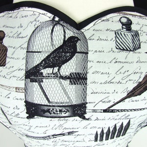 May include: A white fabric with a black and white print of a bird in a cage, a quill pen, and a bottle of ink. The fabric has a handwritten text in French.