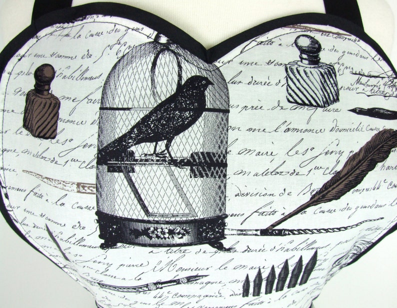May include: A white fabric with a black and white print of a bird in a cage, a quill pen, and a bottle of ink. The fabric has a handwritten text in French.