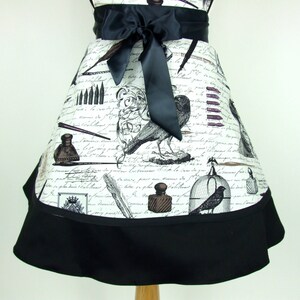 May include: A black and white apron with a vintage design featuring inkwells, pens, and birds. The apron has a black ribbon tie and a black ruffle trim.
