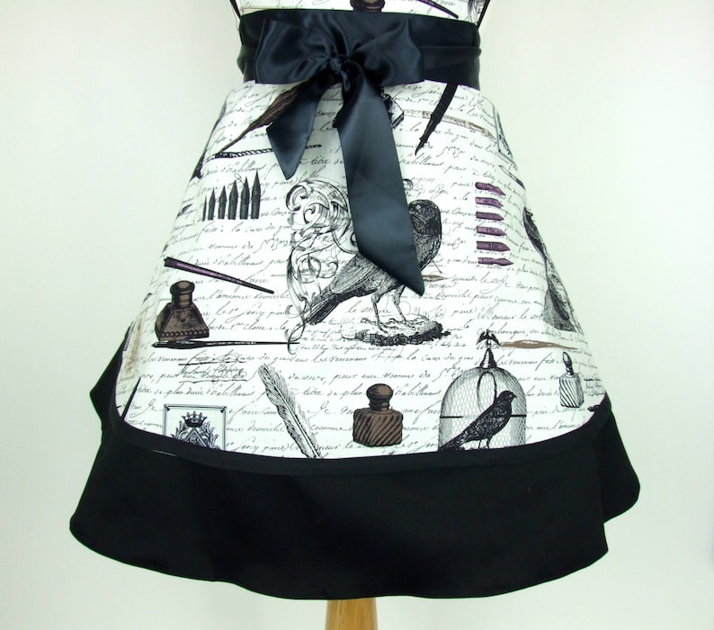 May include: A black and white apron with a vintage design featuring inkwells, pens, and birds. The apron has a black ribbon tie and a black ruffle trim.